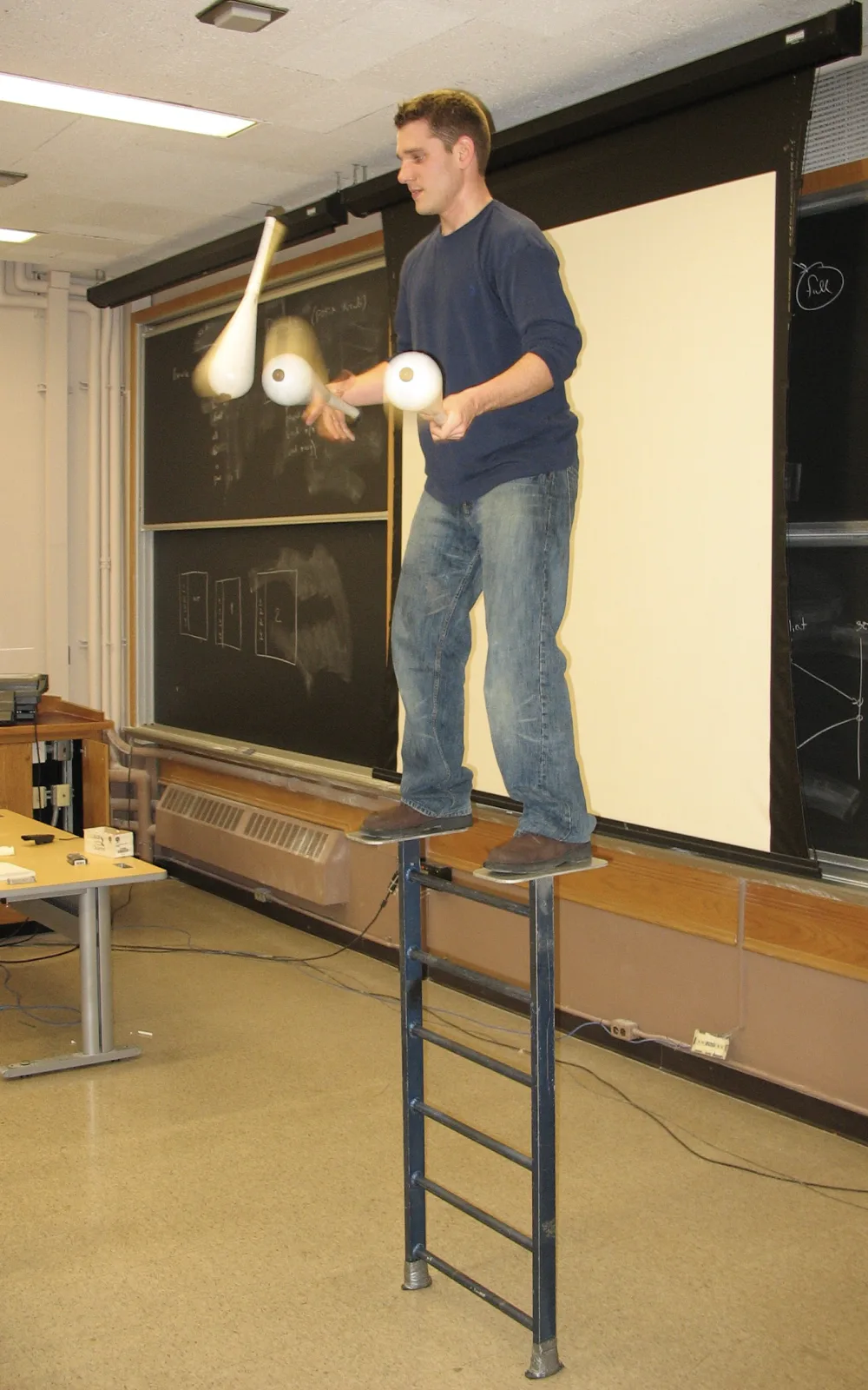 Jacob Abernethy juggling white clubs while standing on two narrow platforms attached to a ladder-like apparatus in a classroom