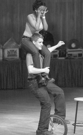 Jacob Abernethy riding a unicycle with a volunteer seated on his shoulders during a comedy juggling performance