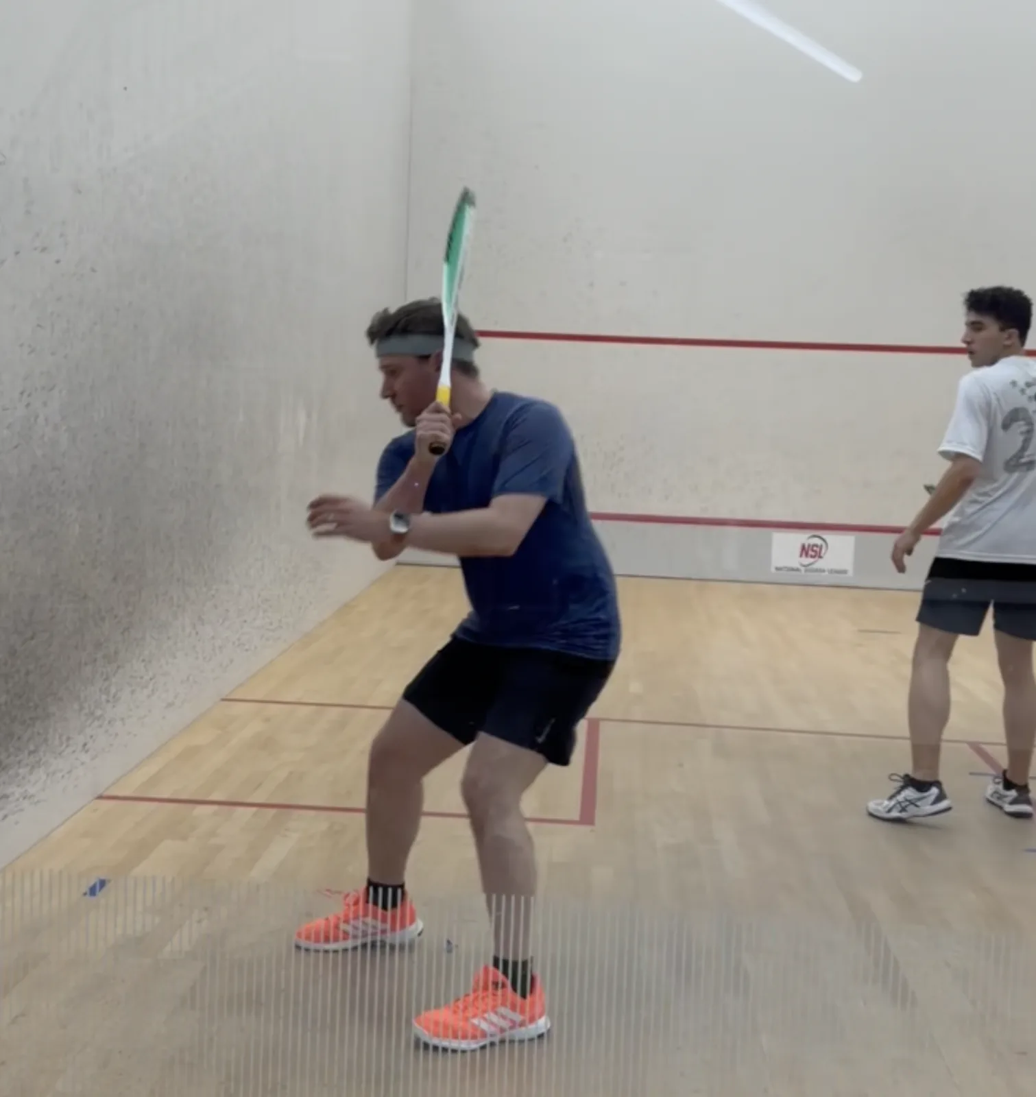 Jacob Abernethy preparing to return a shot on an indoor squash court while another player stands behind him